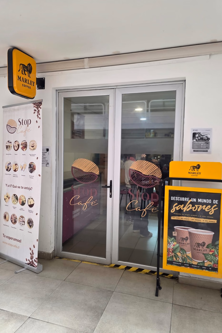 Entrada de Stop Café en ENAC, con la carta en pendón, el logo en las puertas y la señalética de Marley Coffee.